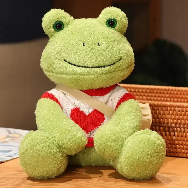 Peluche grenouille avec sa bandoulière et son pull avec un coeur rouge sur une table