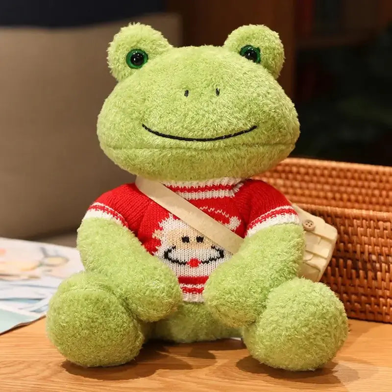 Peluche grenouille avec sa bandoulière et son pull avec un père noël sur une table
