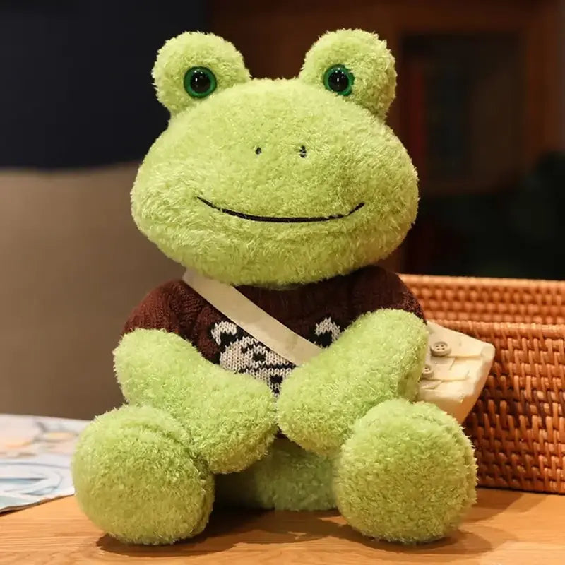 Peluche grenouille avec sa bandoulière et son pull avec un ours brun sur une table