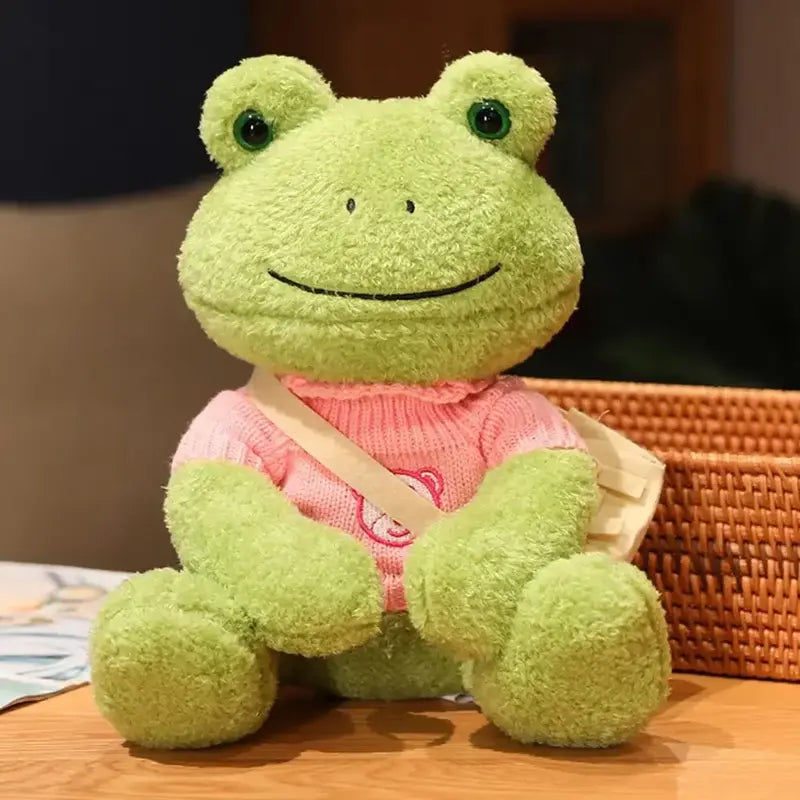 Peluche grenouille avec sa bandoulière et son pull avec ours rose sur une table